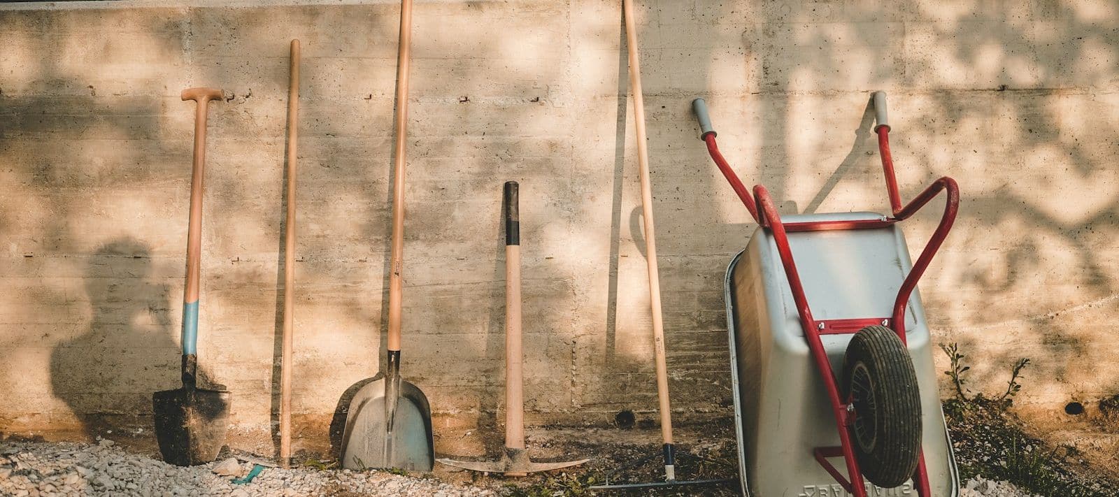 Outils de jardin à manche en bois et brouette appuyés contre un muret ensoleillé, sur du gravier
