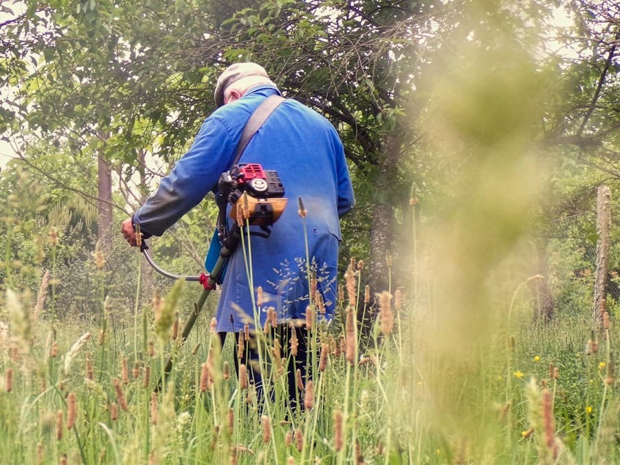 Personne vue de dos, en bleu de travail, passant une débroussailleuse thermique dans l’herbe haute sous les arbres